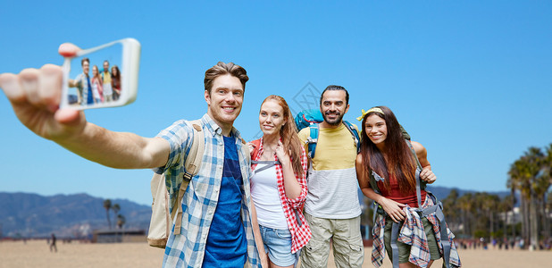 技术,旅游,徒步旅行人们的群微笑的朋友背包加州威尼斯海滩背景上用智能手机自拍背包的朋友用智能手机自拍旅游业高清图片素材