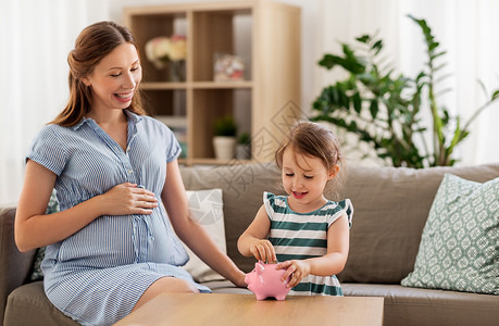怀孕财务家庭观念快乐的怀孕母亲小女儿家里把欧元硬币放进储蓄罐里怀孕的母亲女儿带着储蓄罐小猪高清图片素材