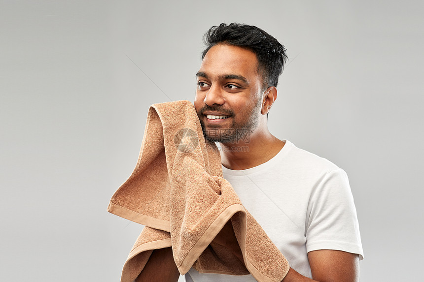 修饰人的微笑的印度男人,用浴巾灰色的背景上擦干他的脸微笑的印度男人带着毛巾灰色的背景上图片
