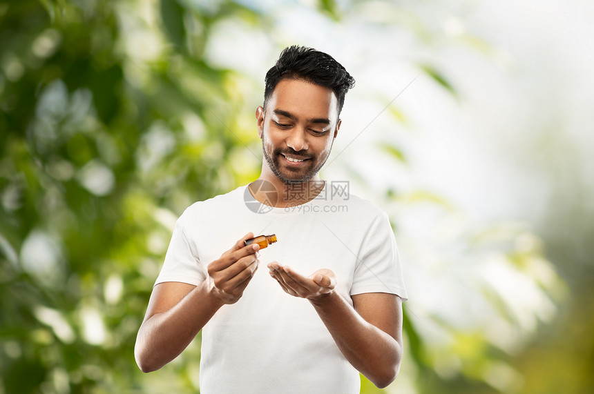 美容,芳香疗法人的微笑的轻印度男子应用精油他的手绿色的自然背景印度男人把美容油涂手上图片