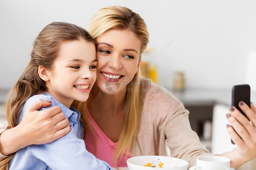 家庭,技术人的快乐的母亲女儿与智能手机吃早餐自拍家里的厨房家人早餐时用智能手机自拍图片