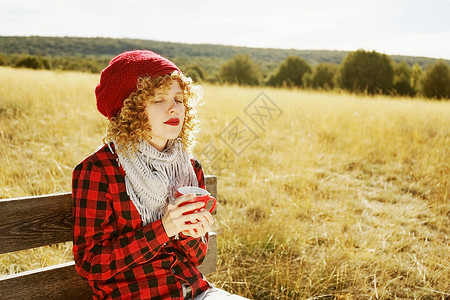 位穿着红色格子衬衫戴着羊毛帽围巾的轻女子的正肖像,她正日光浴,坐黄色田野的木凳上,背光来自秋天的阳光背景图片