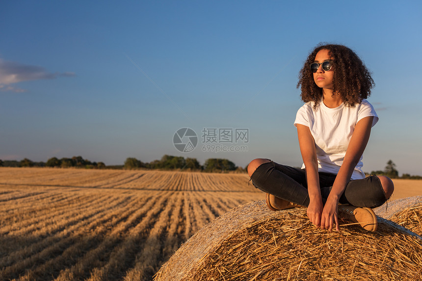 个美丽而体贴的混血非裔美国女女孩,十几岁的轻女人,阳光下戴着太阳镜,坐田野的干草堆上图片