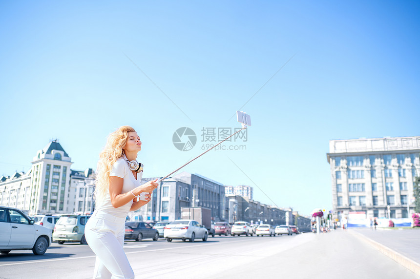 美丽的轻女人戴着音乐耳机脖子上,用自拍棒着城市拍照图片