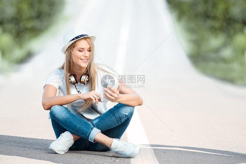 快乐的轻女人,脖子上戴着老式音乐耳机,智能手机上上网,坐路边的分离条上图片