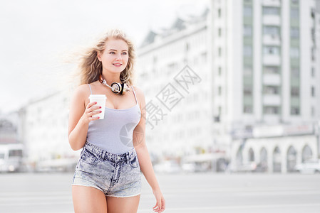 快乐的轻女人带着音乐耳机,着走的咖啡杯,听着音乐,城市背景下行走图片