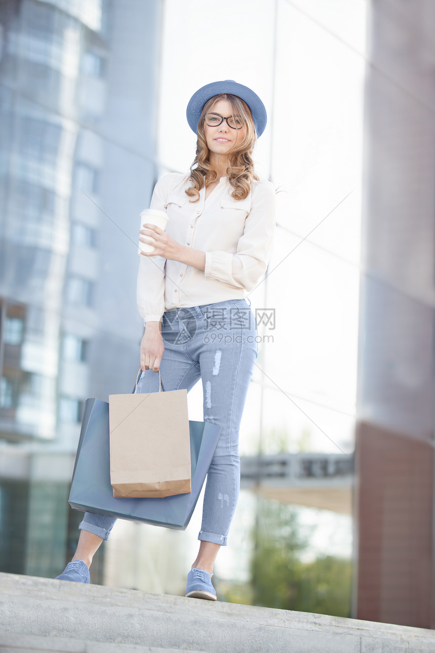快乐的轻时尚女喝着咖啡,站楼梯上,着购物袋,站城市背景下图片