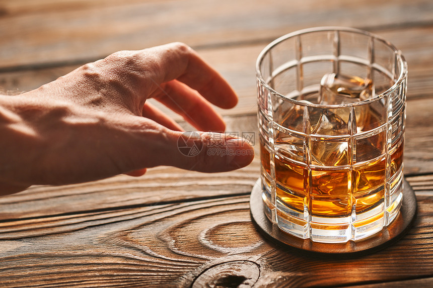 男人的手伸向乡村木桌上的杯加冰块的威士忌酗酒的图片