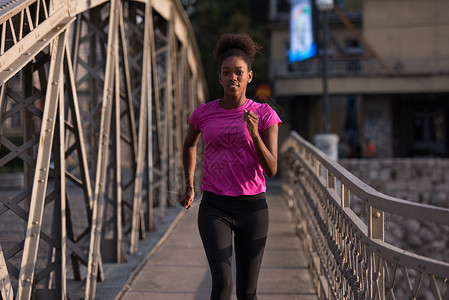 轻运动的非裔美国妇女跑步人行道上过桥,清晨慢跑与城市日出的背景图片