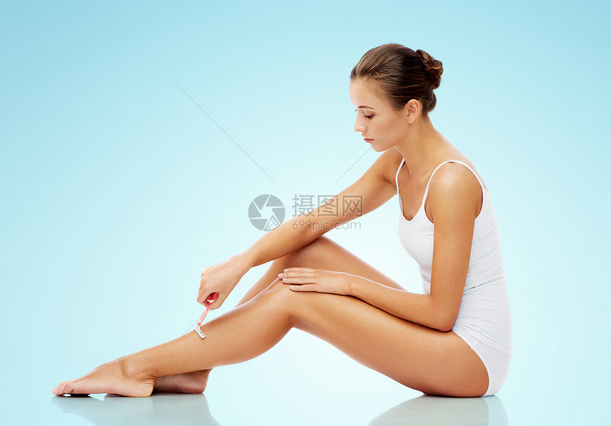 人,美容脱毛美丽的女人与安全剃须刀剃须腿坐地板上的蓝色背景女人带着安全的剃须刀,把腿剃得比蓝色的还要长图片