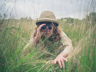 个戴着狩猎帽的轻女人躲高高的草丛里,正用望远镜看双筒望远镜高清图片素材