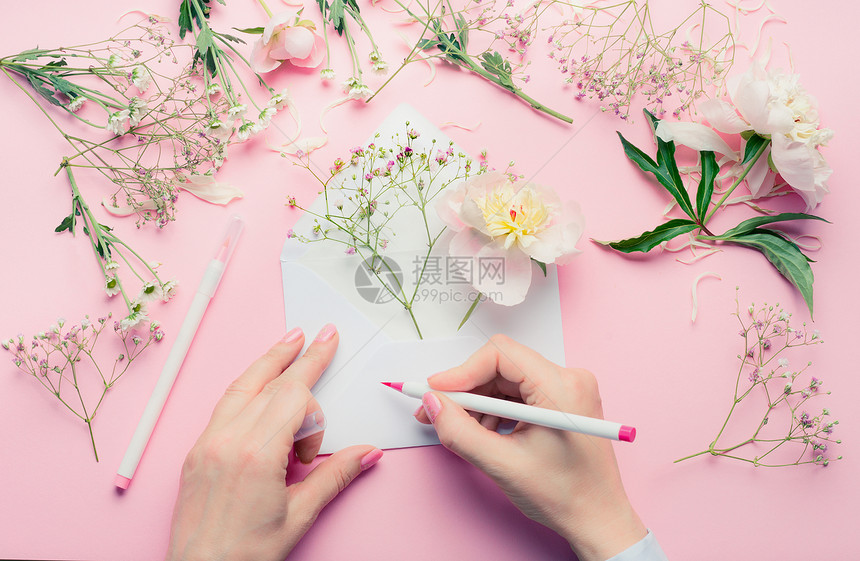 女的手用铅笔写打开的信封上,花的排列花店装饰设备粉红色的桌子背景,顶部视图邀请,问候假期,图片