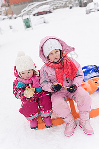 下雪的冬天,两个可爱的小女孩坐户外雪橇上,休息时吃美味的饼干图片