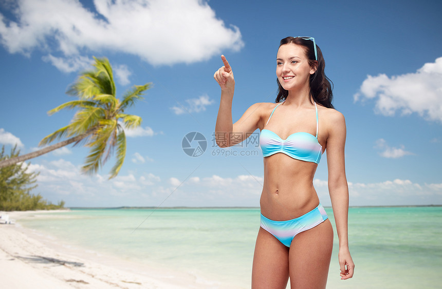 人,旅游,泳装暑假的快乐的轻女人穿着比基尼泳衣指着些想象的东西异国情调的热带海滩与棕榈树的背景图片
