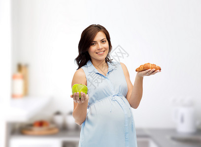 怀孕,健康饮食,垃圾食品人们的快乐的孕妇选择绿色苹果牛角包厨房背景图片