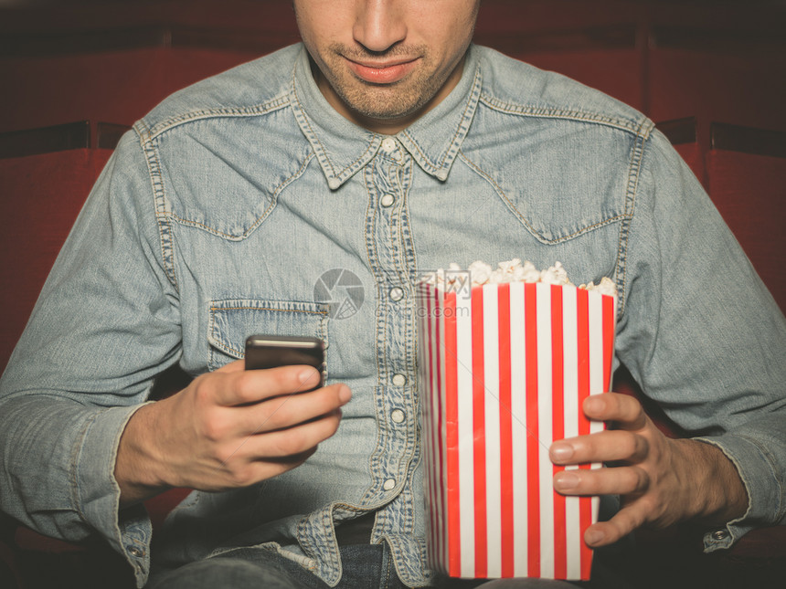 个轻人着桶爆米花坐电影院里,正用他的智能手机图片