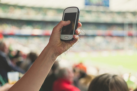 只女的手体育场里着部智能手机,为体育赛事拍照图片