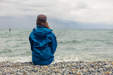 个穿着蓝色夹克的轻女人个刮风的日子里坐海滩上背景图片