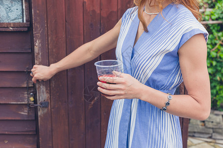 个轻的女人穿着件夏装,着杯饮料,正向个木制的花园小屋开门背景图片