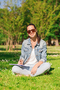 生活方式,暑假,教育人的微笑的轻女孩用铅笔写字笔记本,坐公园的草地上图片