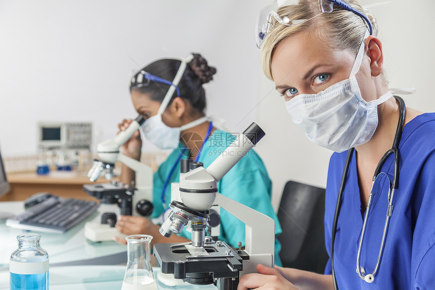个金发的女医学科学研究员医生实验室里用她的显微镜,她的亚洲同事她身后失了注意力图片