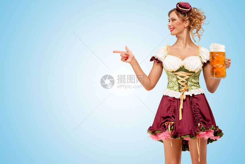 轻感的啤酒节女服务员穿着传统的巴伐利亚连衣裙,着个啤酒杯,指着蓝色的背景图片