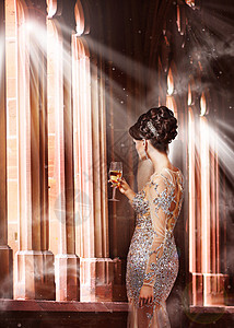 奢侈轻的女人穿着晚礼服,着杯香槟站阳光下的窗户前背景图片