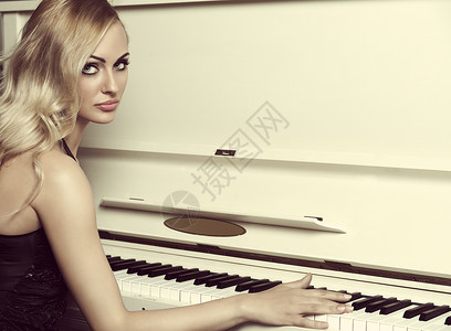 美丽的轻女人,金色的长发,穿着优雅的黑色连衣裙,靠近钢琴图片