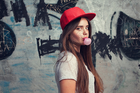 时尚美丽的长发元模特涂鸦背景上摆姿势泡泡糖红色帽子灰色T恤图片