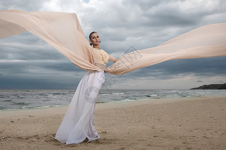穿着米色长裙的女模特海滩上摆出动感十足的造型挥动布料飘动的长裙背景图片