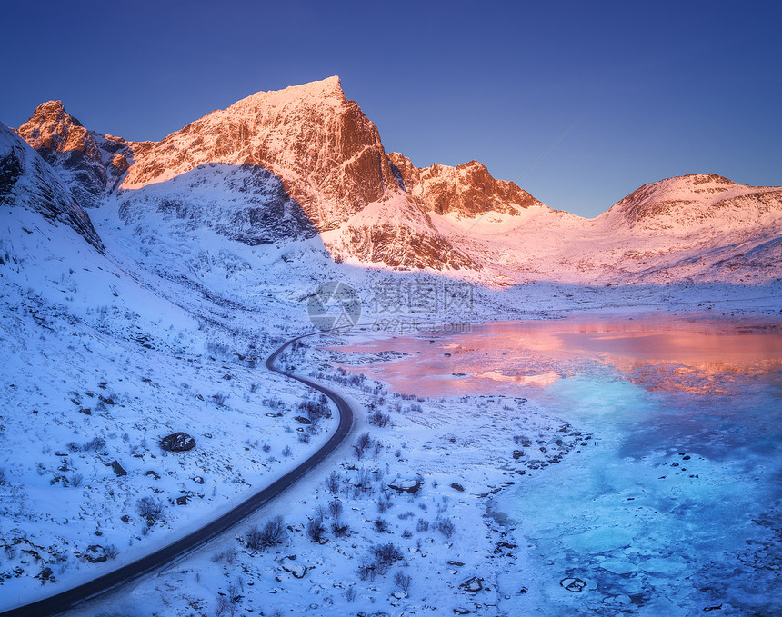 鸟瞰蜿蜒的道路,积雪覆盖的山脉蓝天反射水日落冬季景观与海洋,霜冻海岸,雪岩,道路晚上挪威的洛芬岛图片