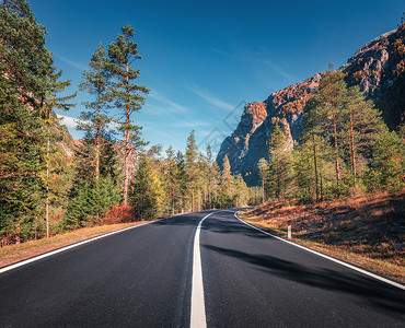 意大利日落时秋天森林的道路美丽的山路,绿色的树,橙色的草,高高的岩石,蓝天景观与空沥青路穿过森林秋天旅行背景背景图片
