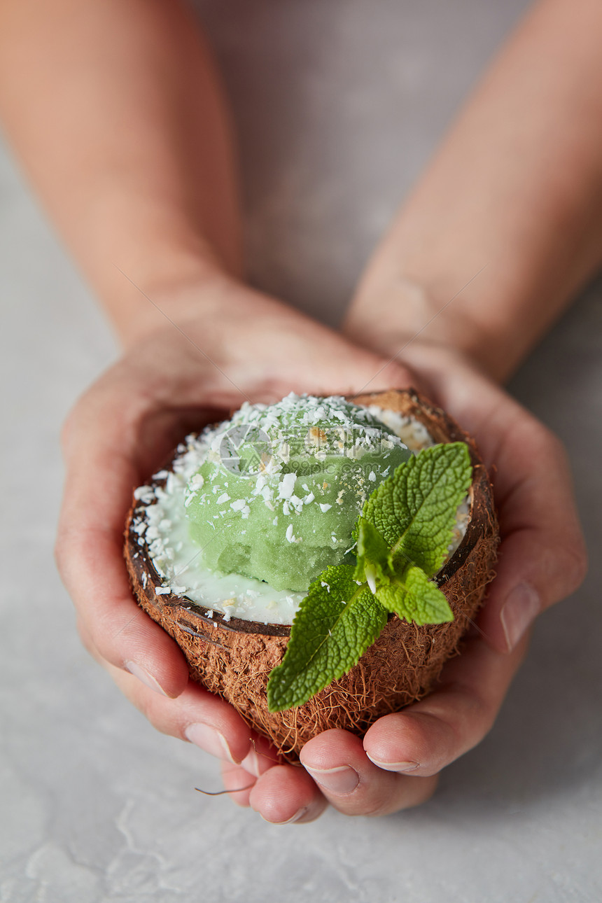 薄荷绿色冰淇淋自制的椰子壳女人手上的灰色混凝土背景素食者的夏季甜点女人的手着冷冻的绿色冰糕椰子壳与新鲜的薄荷叶灰色图片