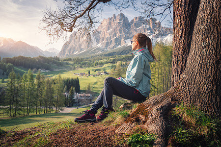 日落时,戴着太阳镜蓝色夹克的漂亮轻女人坐树下的山上五颜六色的春天景观与运动女孩,草地,绿树山脉意大利旅行背景图片
