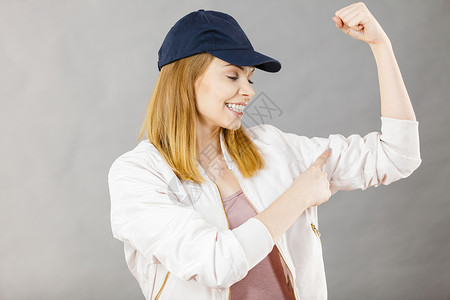 轻的运动女戴着帽子运动服,展示她的手臂肌肉享受锻炼结果工作室拍摄灰色背景轻的女人展示她的手臂肌肉背景图片