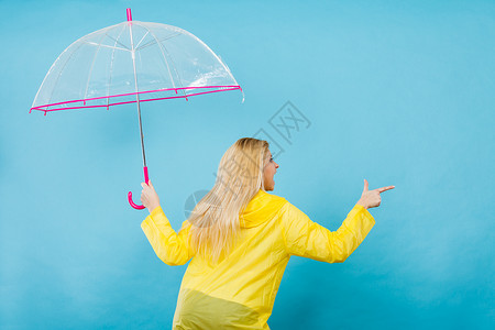 金发女人穿着黄色雨衣,着透明的雨伞,检查天气否下雨,用手指指着电脑图片