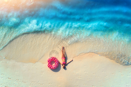 鸟瞰美丽的轻躺着的女人,粉红色的甜甜圈游泳环白色的沙滩上,靠近大海,日落时波浪暑假苗条女孩的最高视野,清澈的蔚蓝的背景图片