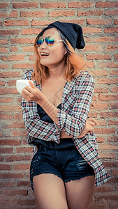 快乐的年轻时髦女人站在街上砖墙旁边手里拿着咖啡杯杯子高清图片素材