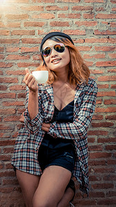 快乐的年轻时髦女人站在街上砖墙旁边手里拿着咖啡杯漂亮的高清图片素材