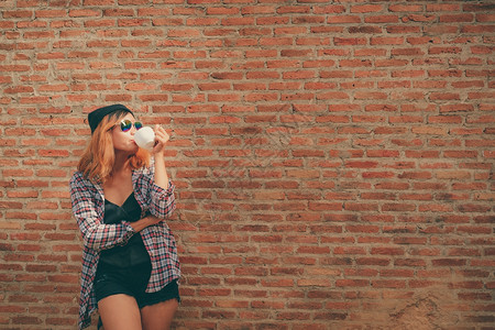 快乐的年轻时髦女人站在街上砖墙旁边手里拿着咖啡杯帽子高清图片素材