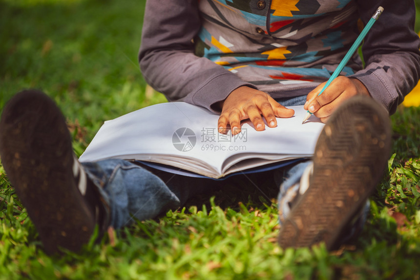 小孩坐在公园草地上用铅笔在笔记本上写作图片