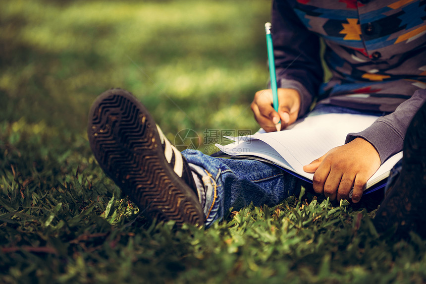 小孩在公园绿草地上用铅笔在笔记本上写作图片