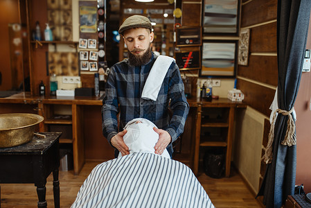 理发师和顾客用毛巾擦脸剪胡子专业理发店是一种时髦的职业男美发师和客户在发廊剃须前的程序理发师和顾客脸上带着毛巾理发店切割高清图片素材