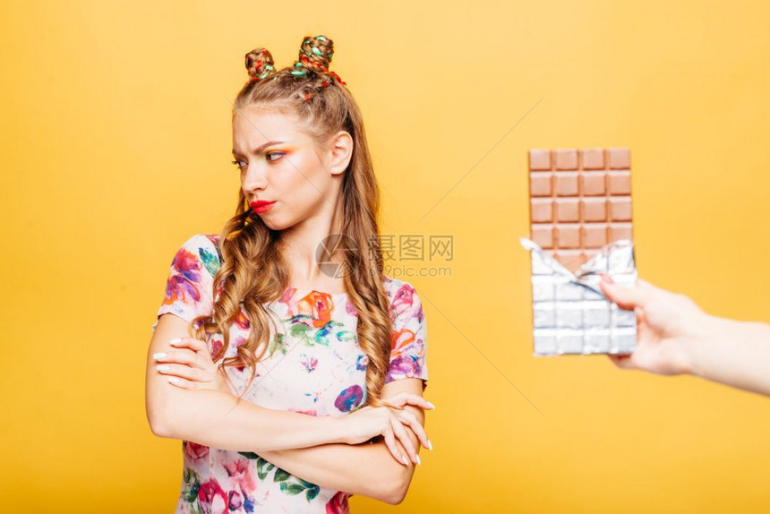 美丽的年轻女士拒绝吃巧克力吧穿着金色卷发的明亮女孩穿着夏季彩色服装的时髦女孩背景上是黄色的墙女士拒绝吃巧克力吧图片
