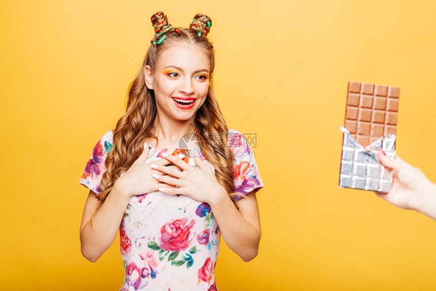 美丽的年轻女士惊讶地看到有人给了她大巧克力棒光亮的金发女孩时髦的穿着夏季多彩服装的女孩背景上黄色墙女人惊讶地看到有给了她大巧克力图片