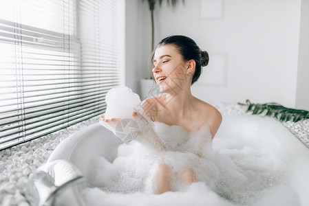 浴室泡沫坐在浴缸里玩泡沫的笑着女人豪华的浴室有窗户和棕榈树枝装饰背景