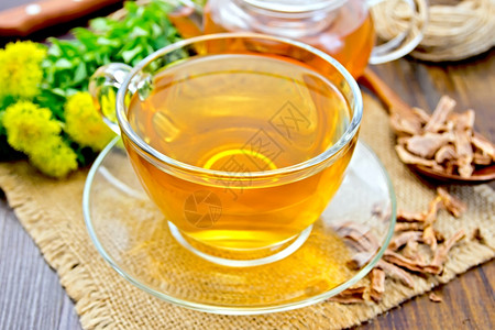 一杯玻璃中的草药茶和一块薄饼餐巾上的茶壶有干根勺子和木板背景上的鲜花罗迪奥拉玫瑰自然高清图片素材