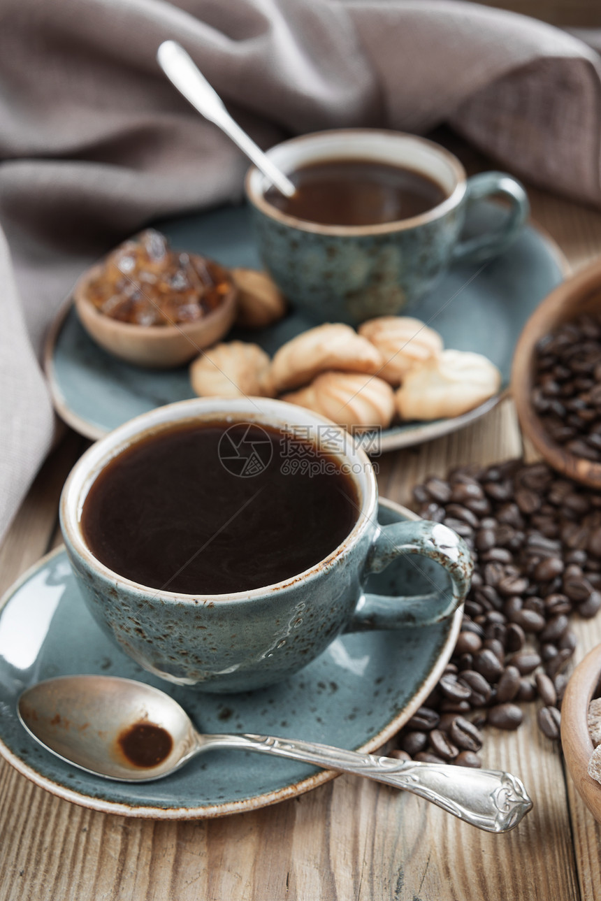 两杯黑色咖啡饼干和糖的蓝杯黑咖啡饼干和糖片由旧木制餐桌上的布环绕图片