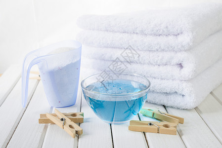 3条白色毛浴巾测量杯中的洗衣粉玻璃碗中的蓝织布软体和白板背景上的木织物背景图片
