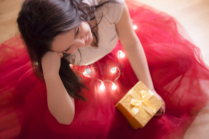 身着红色礼服坐在地板上四周都是圣诞灯光并拿着一个装捆绑弓的礼品盒图片
